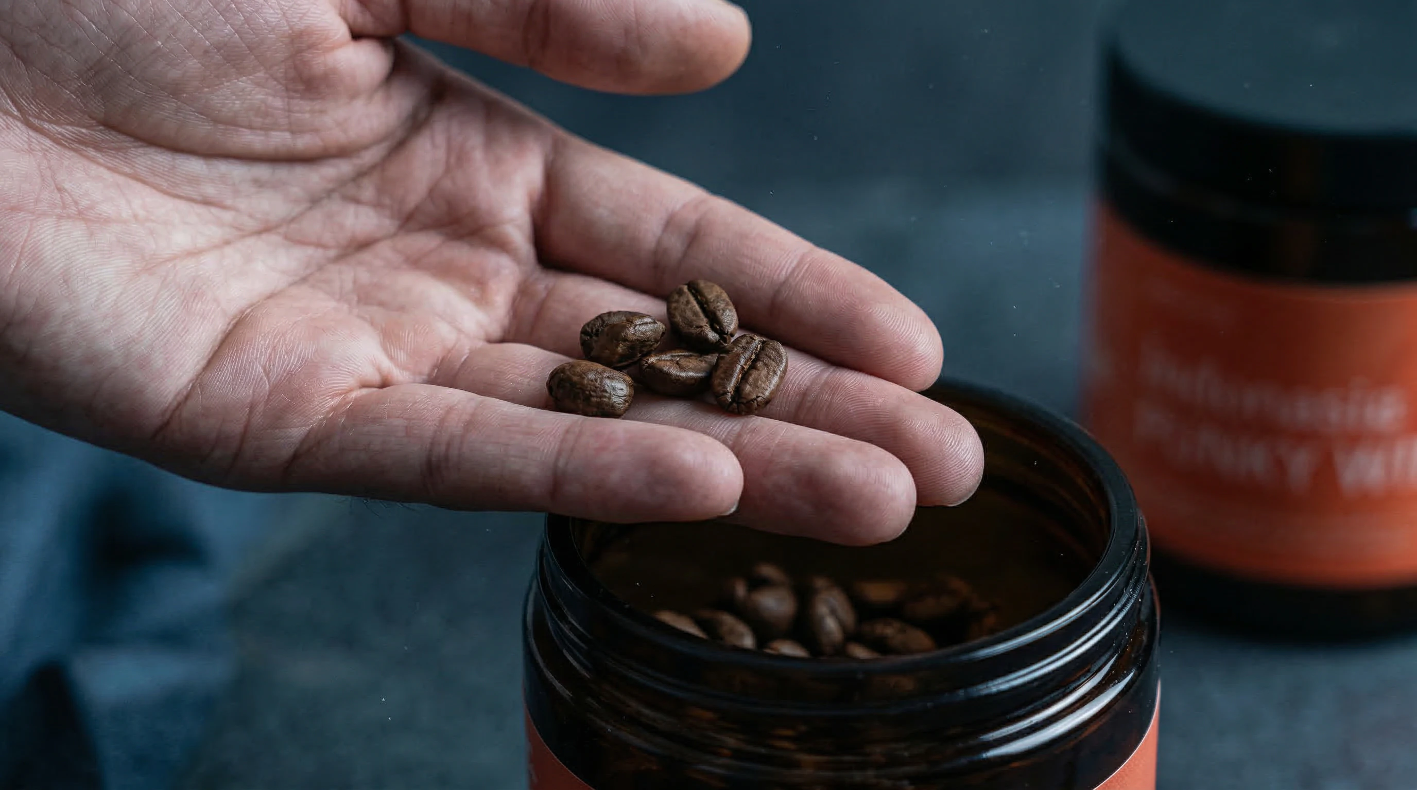 Hand holding roasted coffee beans above an open jar in moody blue tones